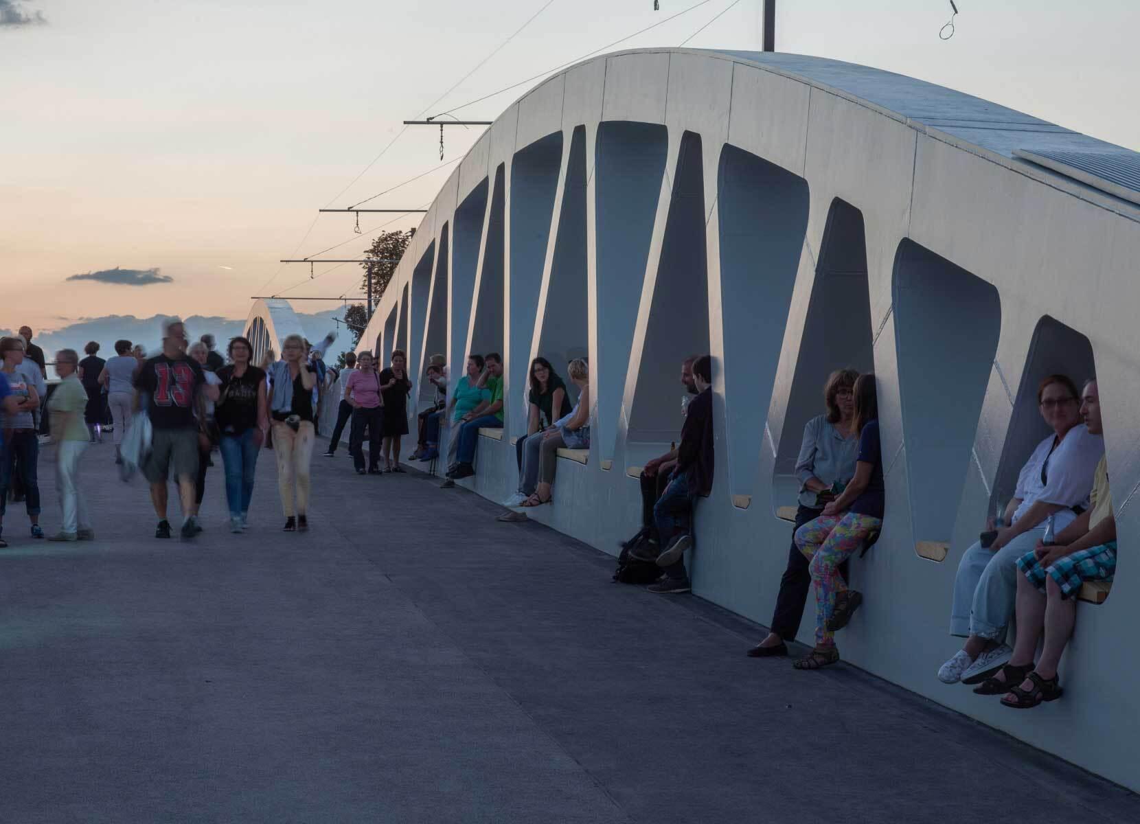 Des usagers franchissant un pont contemporain et utilisant les ouvertures dans la poutre pour s’asseoir